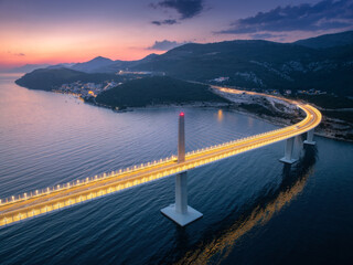 Aerial view of modern Peljesac bridge and blue sea at night. Croatia. Bridge with illumination, mountains, reflection and purple sky at sunset. Top drone panoramic view of road, street lights at dusk