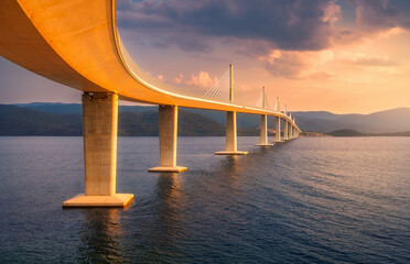 Modern Peljesac bridge and blue sea at colorful sunset in summer. Croatia. Beautiful Peljeski bridge during golden hour, mountains, reflection in water, orange sky with clouds. Architecture. Landscape