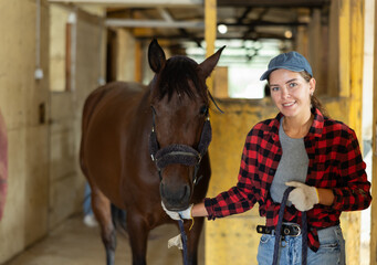 Positive young girl in plaid shirt and gloves takes horse out of stall at big stable