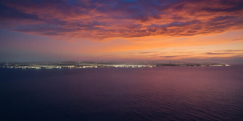 Vibrant purple and orange sunset sky over calm sea with distant illuminated coastline

