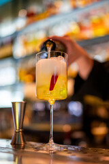 Bartender pouring a foamy cocktail into a tall glass with red garnish and olives on a blurred bar background. Concept of nightlife, mixology, and professional drink service.