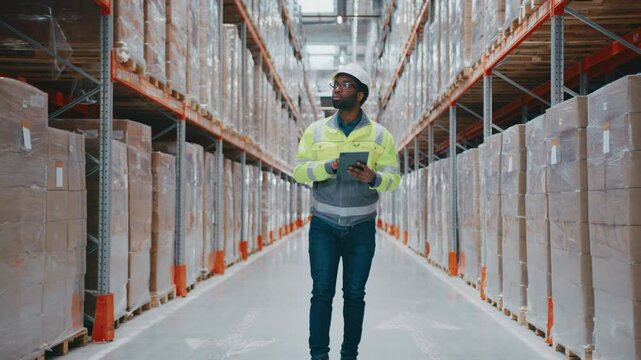 Attentive male worker in helmet holding tablet and walking between storage racks. Looking around and inspecting shelves. Monitoring stock levels or reviewing warehouse layout digitally.