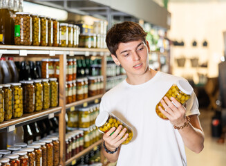 Young guy in supermarket selects examines compares two cans of olives. Male customer chooses jar of...
