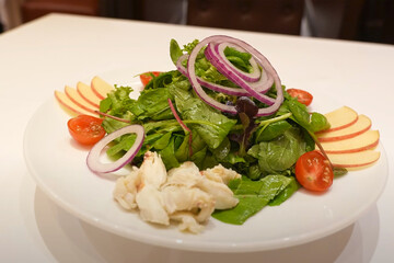 Fresh mixed green salad served with red onion rings, cherry tomatoes, apple slices, and lump crab meat on a white plate in a fine dining setup

