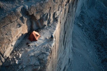 A climber prepares a tent on a narrow ledge high above the ground as the sun rises, illuminating the rocky landscape and mountains