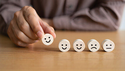 Hand selecting a happy face among feedback emoticon blocks on a wooden table showcasing customer satisfaction and positive reviews.