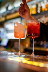 Bartender stirring cocktail in tall glass with metal stirrer at a bar. Blurred background, vibrant nightlife atmosphere, focus on drink preparation and mixology.
