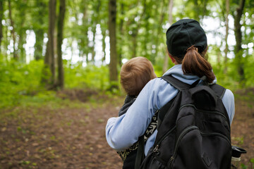 Rear view of mother carrying baby in carrier during walk through forest. Woman with backpack hiking in green woodland, enjoying outdoor family time.