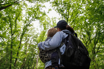
Mother carrying baby in baby carrier during forest trip. Woman with backpack walking through green forest enjoying time outdoors with family.