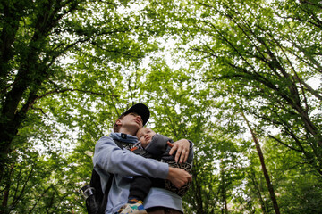Mother holding baby in arms during walk in forest. Woman with backpack walking in green forest enjoying time outdoors with family.