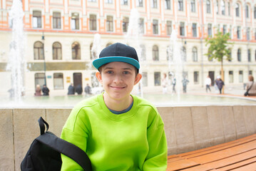 A 12 year old boy in a cap and light green clothes sits on a bench near a fountain on the Stock...