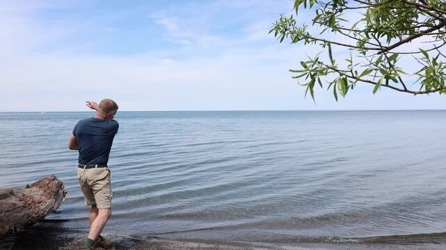 Middle-aged man in a blue shirt skips stones into a smooth blue lake