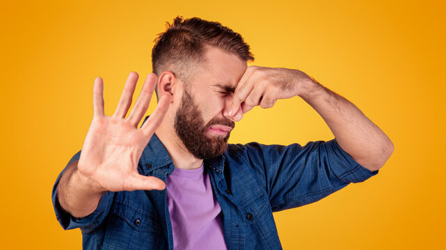 Terrible smell concept. Millennial hipster guy closing his nose and gesturing STOP, feeling disgusted on grey studio background. Young man repulsed by unpleasant odor or stinky perfume
