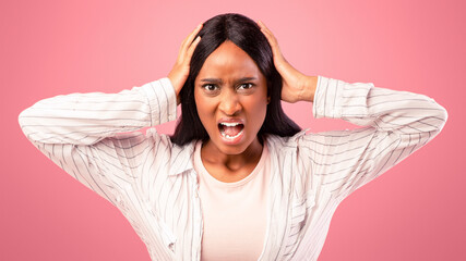 Furious black woman screaming and holding her head on pink studio background. Emotional African...