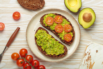 Slices of whole grain rye bread with mashed avocado, green peas and cherry tomatoes seasoned with black sesame seeds are served on a plate for a healthy breakfast or snack