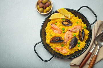 Seafood paella with rice, shrimp, mussels, peas, and lemon wedges served in a black traditional pan on a grey rustic background with olives and cutlery, copy space