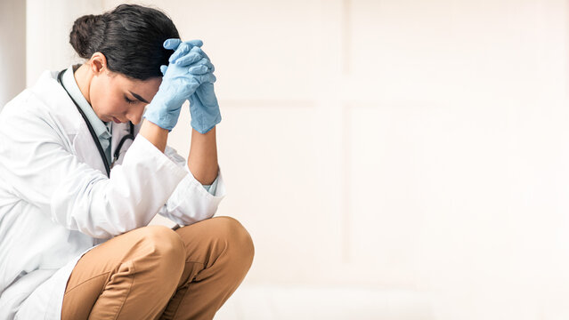 Stressed brunette young woman doctor having difficult day at clinic, wearing protective gloves, sitting next to wall in her office and resting, empty space. Upset female doctor feeling exhausted