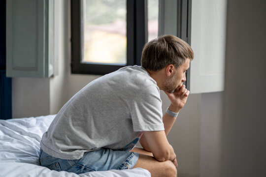 Thoughtful lonely man sitting on bed by window lost in deep thought. Reflective melancholy male feeling uncertain, emotional fatigue, overwhelmed with personal problems. Pensive mood, mental burnout.