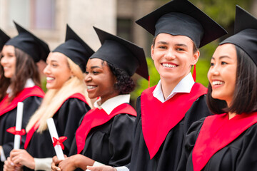 Cheerful handsome guy standing among international group of students, wearing graduation costumes...