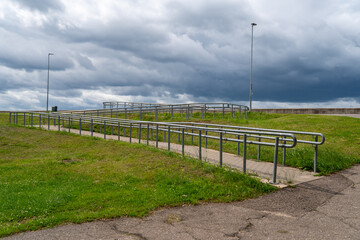 concrete wheelchair accessible ramp with metal railings on grassy slope under dramatic cloudy sky,...