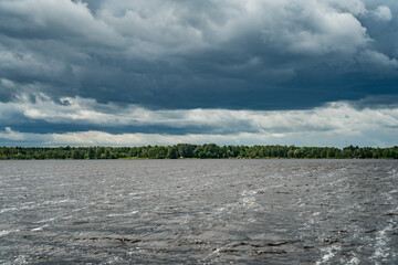 stormy river Daugava with dark dramatic clouds above choppy water and distant forest shoreline, natural weather landscape, moody sky over water surface, scenic outdoor climate and storm atmosphere