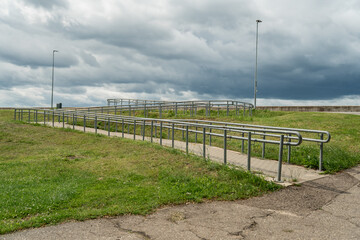 concrete wheelchair accessible ramp with metal railings on grassy slope under dramatic cloudy sky,...