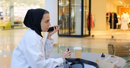 Young muslim woman wearing hijab is sitting at a table in a shopping mall, recording voice notes on her phone and taking notes on her tablet. Free and modern Muslim in city