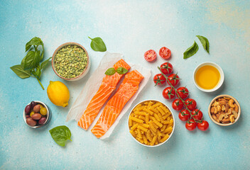 Raw salmon fillets, pasta, cherry tomatoes, olives, nuts, peas, lemon, and basil arranged on a blue...