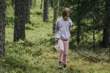 A young girl in a lavender hoodie and pink pants walks through a mossy forest, holding a plastic jar, possibly collecting insects or exploring nature.