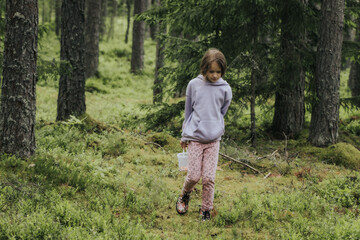 A young girl in a lavender hoodie and pink pants walks through a mossy forest, holding a plastic jar, possibly collecting insects or exploring nature.
