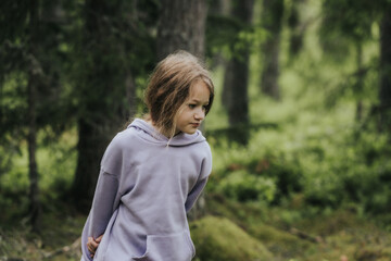 A young girl in a lavender hoodie and pink pants walks through a mossy forest, holding a plastic jar, possibly collecting insects or exploring nature.