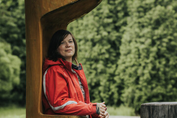 A woman in a red outdoor jacket leans against a large wooden structure with a calm expression, surrounded by a lush green forest background.