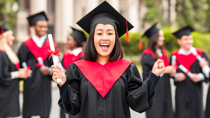 Delighted asian young lady in graduation costume showing her diploma and screaming at camera, happy...