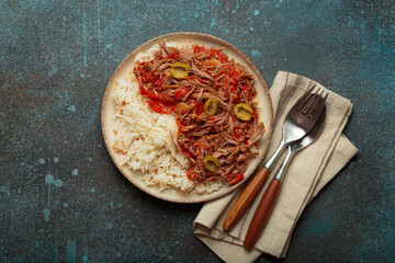 Ropa vieja, a traditional Cuban dish, is served with white rice, tomato sauce, and green olives on a plate from above on dark rustic background