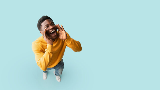 Cheerful african american guy posing over blue studio background, holding hands to mouth and saying something out loud, panorama with copy space. Handsome young black man making announcement, top view