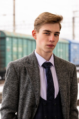 A young man in formal vintage clothing stands at a train station with a serious expression
