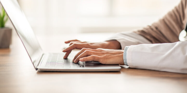 Cropped of woman doctor having online training, sitting in her cabinet, using laptop, typing on keyboard, side view, copy space. Unrecognizable lady physician working with computer, clinic interior