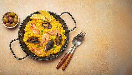 Seafood paella with rice, shrimp, mussels, peas, and lemon wedges served in a black traditional pan on a light beige background with olives and cutlery