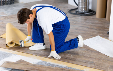 Skilled young carpenter using rubber mallet to lay click-lock laminate flooring during interior...