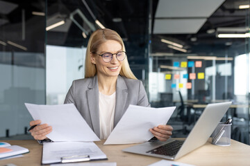 A smiling businesswoman reviews documents at her desk in a modern office setting, alongside a...