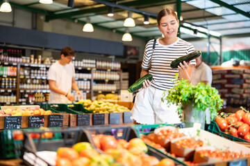 Smiling young girl shopping for organic vegetables in farm store, choosing ripe green zucchini in boxes of produce display
