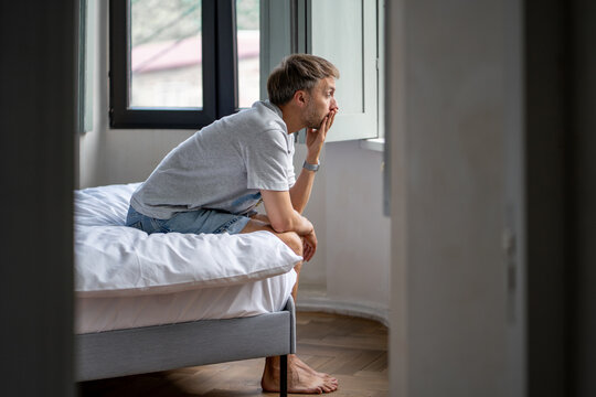Tired man sits on bed lost in deep thought after sleepless night. Exhausted male suffering from insomnia, feeling lonely, overthinking problems, dealing with relationship breakup, midlife challenges.