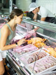 Young woman buyer of meat department of grocery store, looking at raw meat small sausage in...