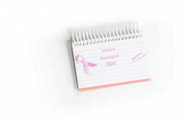 Schedule mammogram today message written on a lined index card notebook with a pink breast cancer awareness ribbon and paper clip, white background.
