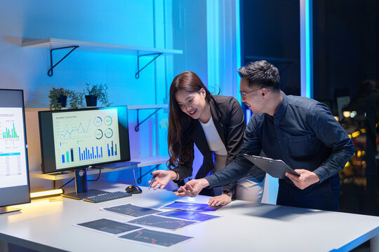 
Young Asian business team working late into the night in a well-lit office, collaborating on a project using laptops.