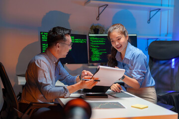 Two Asian software engineers collaborate at night in modern office, writing machine learning algorithms and debugging artificial intelligence code on multiple monitors in AI tech development workflow.