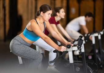 In gym, girl does exercises using exercise bicycle. Young group of athletes trains in gym. Physical...