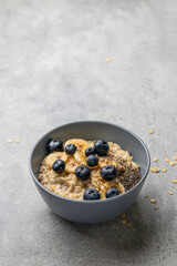 Oatmeal porridge in bowl with milk, blueberries, banana, chia seeds and honey on a grey background. Healthy breakfast homemade food. Home cooking. Selective focus, copy space.