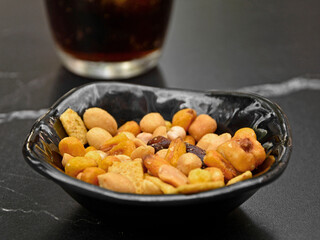 orzeszki w czarnej miseczce na stole obok szkalnki z napojem, przekąska z orzeszkami, miks orzeszków jako przekąska, mix nuts in a bowl as a snack
