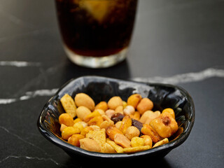 orzeszki w czarnej miseczce na stole obok szkalnki z napojem, przekąska z orzeszkami, miks orzeszków jako przekąska, mix nuts in a bowl as a snack
